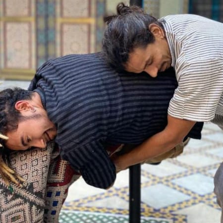 two performers train in a indoor rehearsal studio laid with patterned carpets, one performer is embracing the other resting their head on their back, the other performer holds a rolled up carpet mimicking the embrace, both have facial hair and wear striped shirts and dark jeans Zarbia | Company Zid and Brahim Ech-Chibi Zarbia work-in-progress