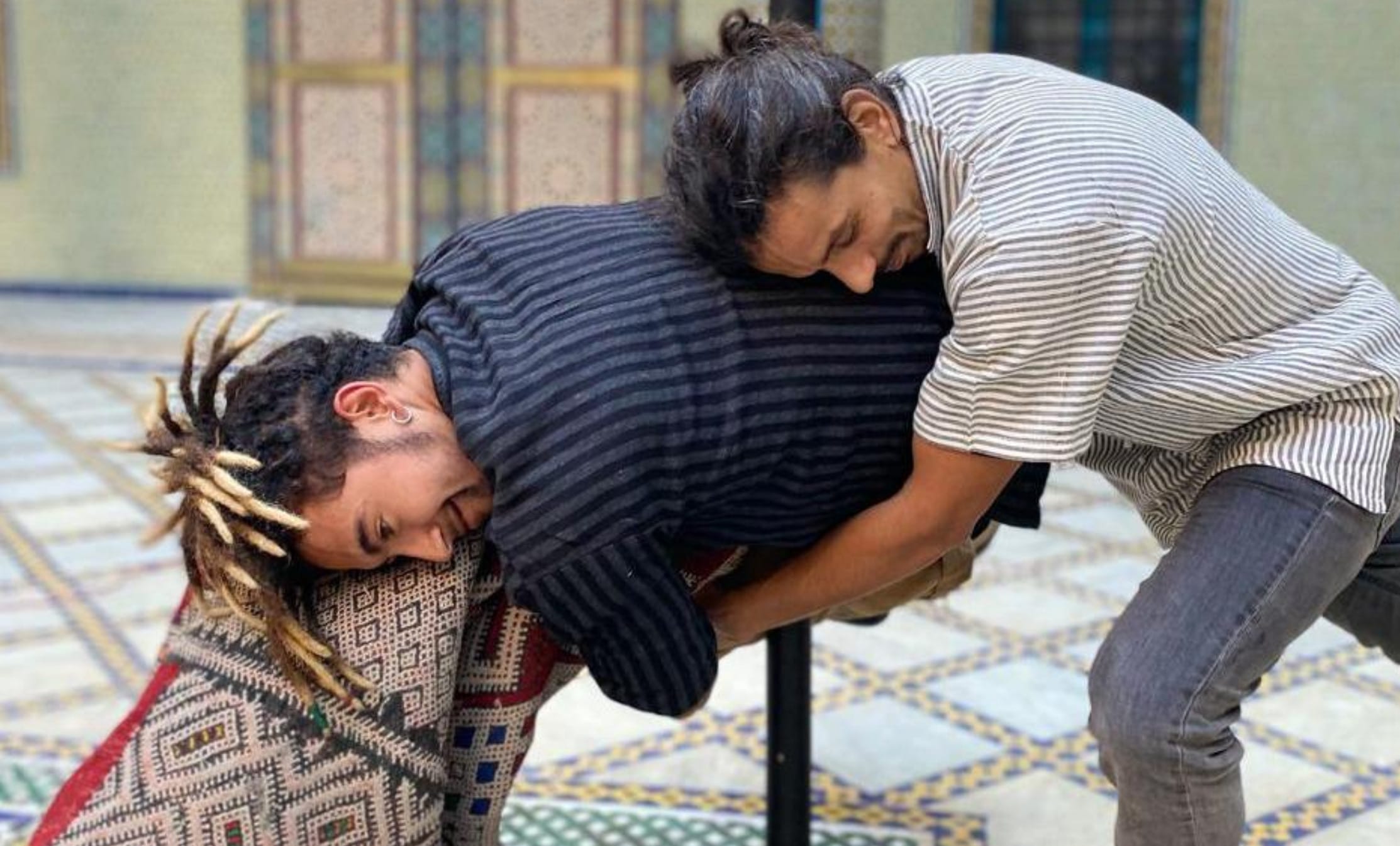 two performers train in a indoor rehearsal studio laid with patterned carpets, one performer is embracing the other resting their head on their back, the other performer holds a rolled up carpet mimicking the embrace, both have facial hair and wear striped shirts and dark jeans Zarbia | Company Zid and Brahim Ech-Chibi Zarbia work-in-progress