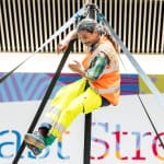 a performer in an indoor busy brightly lit shopping centre performs on a secured Chinese pole, with dark hair in a pony tail and wearing a hi-vis orange vest, yellow hi-vis trousers and green socks, they smile engagingly at the audience Taroo | Company Zid (c) Jimmy Lee