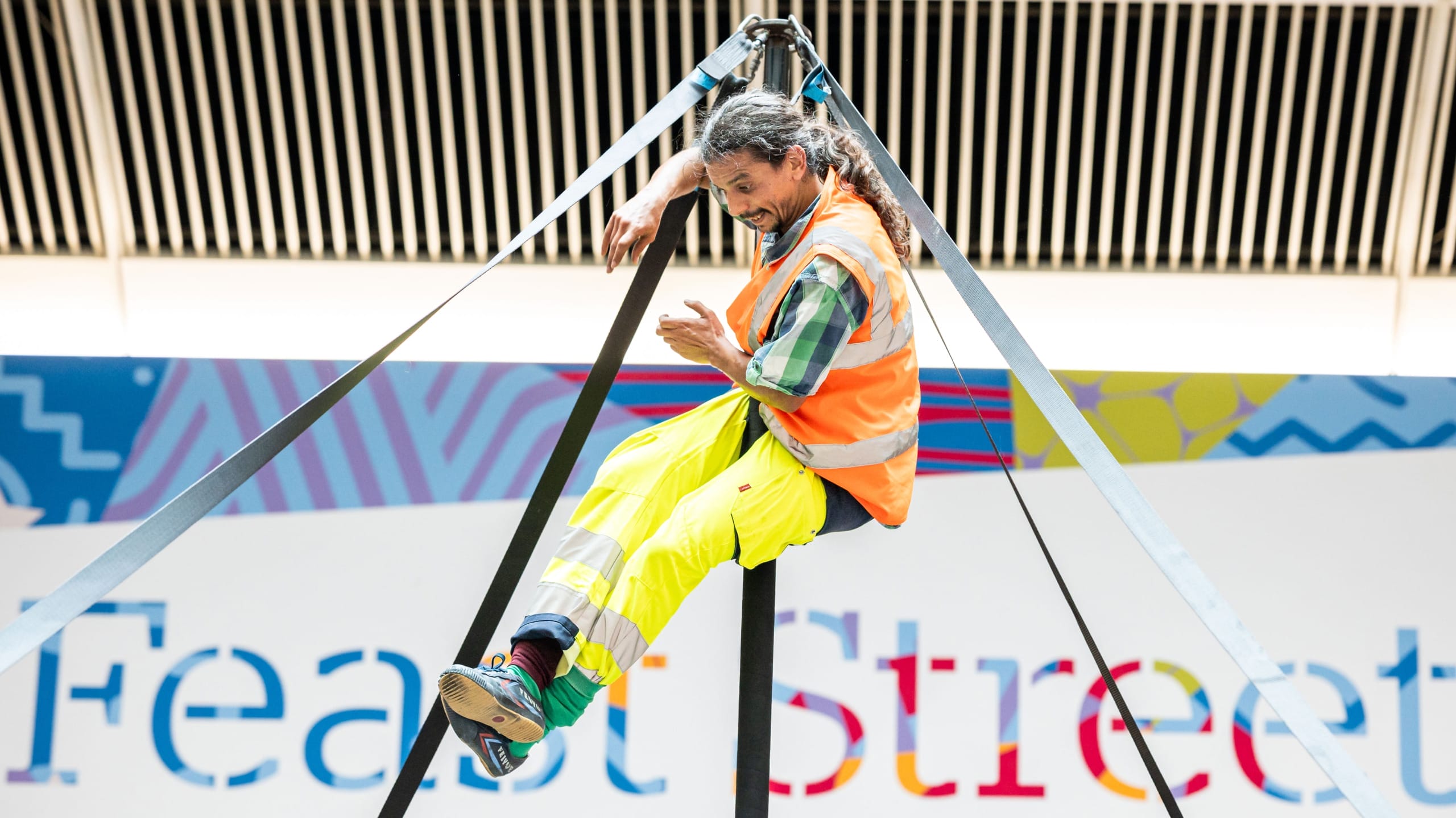 a performer in an indoor busy brightly lit shopping centre performs on a secured Chinese pole, with dark hair in a pony tail and wearing a hi-vis orange vest, yellow hi-vis trousers and green socks, they smile engagingly at the audience Taroo | Company Zid (c) Jimmy Lee