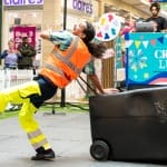 a performer in an indoor busy brightly lit shopping centre performs by a black household bin, with dark hair in a pony tail and wearing a hi-vis orange vest and yellow hi-vis trousers, they smile engagingly at the audience Taroo | Company Zid (c) Jimmy Lee