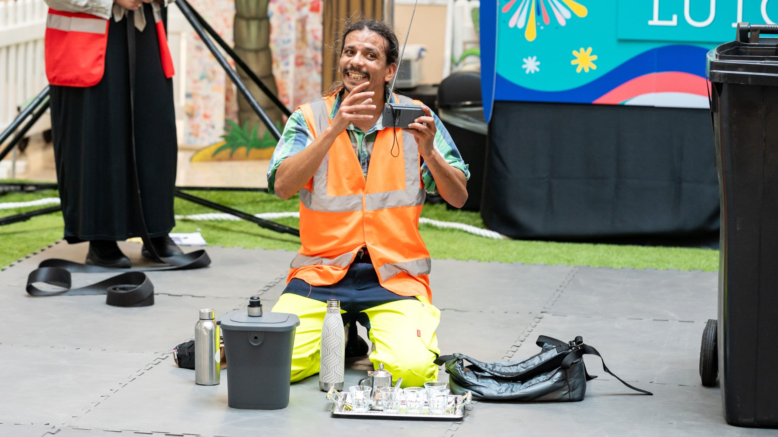 a performer in an indoor busy brightly lit shopping centre performs by a black household bin, a portable radio and a tea set, wearing a hi-vis orange vest and yellow hi-vis trousers, they smile engagingly at the audience Taroo | Company Zid (c) Jimmy Lee