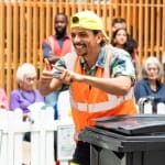 a performer in an indoor busy brightly lit shopping centre performs by a black household bin, wearing a hi-vis orange vest and a yellow backwards baseball cap, they smile engagingly at the audience Taroo | Company Zid (c) Jimmy Lee