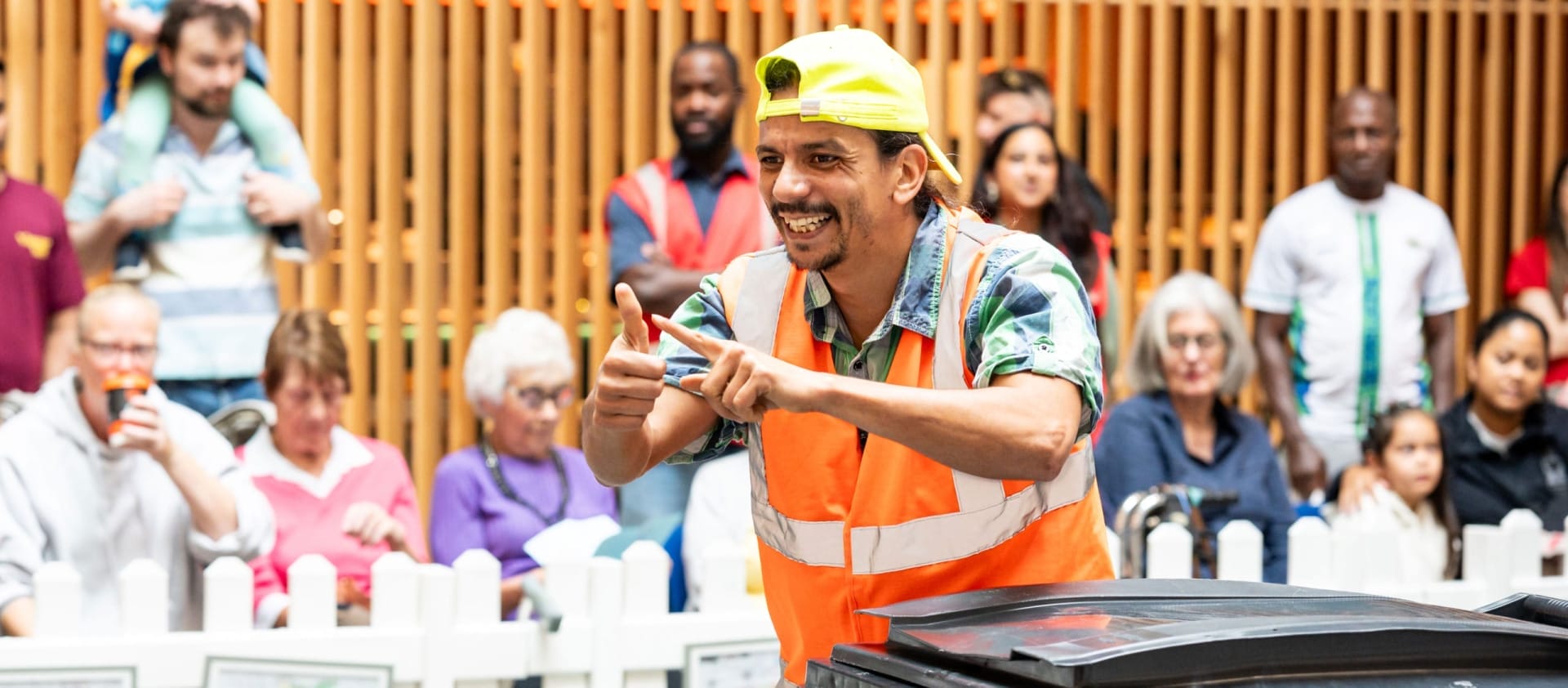 a performer in an indoor busy brightly lit shopping centre performs by a black household bin, wearing a hi-vis orange vest and a yellow backwards baseball cap, they smile engagingly at the audience Taroo | Company Zid (c) Jimmy Lee