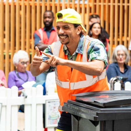 a performer in an indoor busy brightly lit shopping centre performs by a black household bin, wearing a hi-vis orange vest and a yellow backwards baseball cap, they smile engagingly at the audience Taroo | Company Zid (c) Jimmy Lee