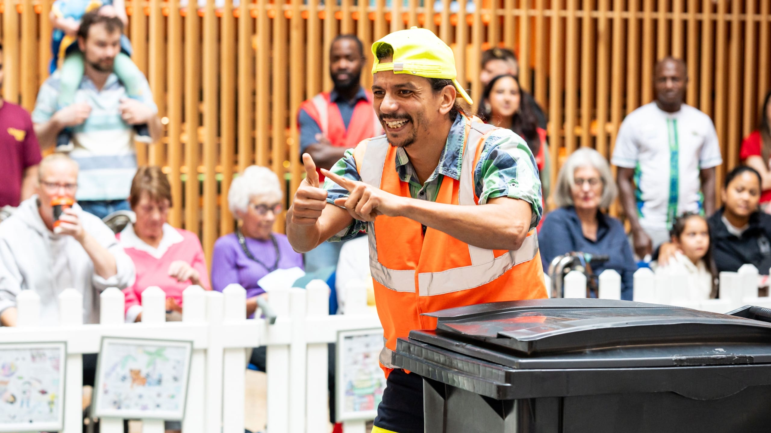 a performer in an indoor busy brightly lit shopping centre performs by a black household bin, wearing a hi-vis orange vest and a yellow backwards baseball cap, they smile engagingly at the audience Taroo | Company Zid (c) Jimmy Lee