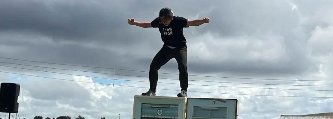 non binary East Asian performer stands on top of a large open fridge, wearing black jeans and t-shirt, holding their arms out against a grey sky I Am. Am I | Louiseanne Wong | MBMS festival FATC story dance circus voice