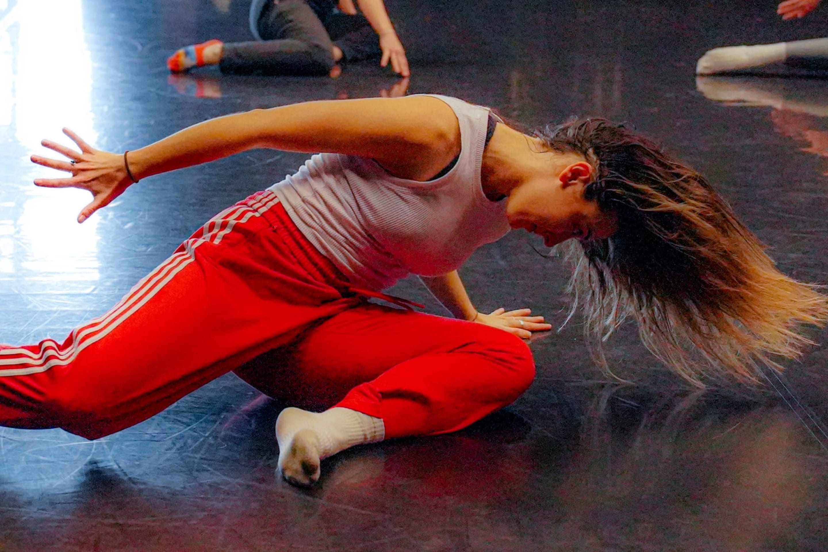 dancer wearing a white vest and red Adidas tracksuit bottoms and white socks moves on the floor of an indoor dance studio