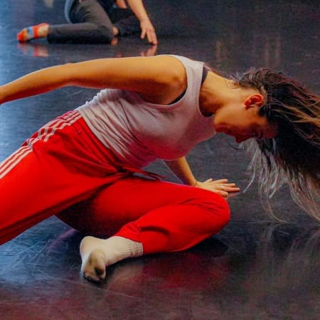dancer wearing a white vest and red Adidas tracksuit bottoms and white socks moves on the floor of an indoor dance studio