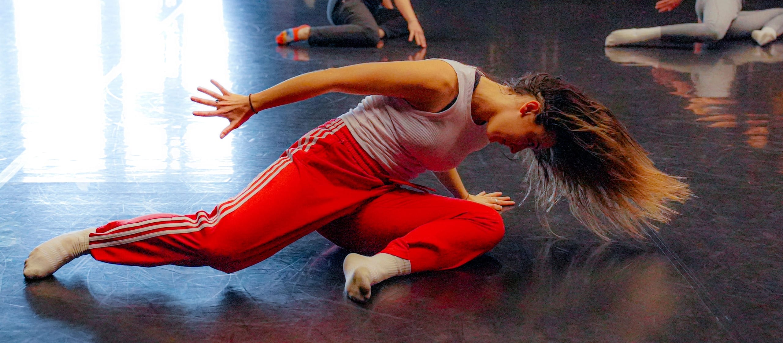 dancer wearing a white vest and red Adidas tracksuit bottoms and white socks moves on the floor of an indoor dance studio